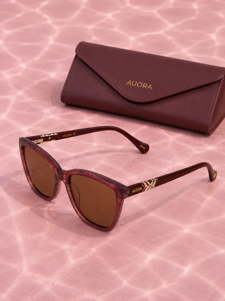 Auora Eyewear burgundy sunglasses with a matching premium case on a pink reflective background